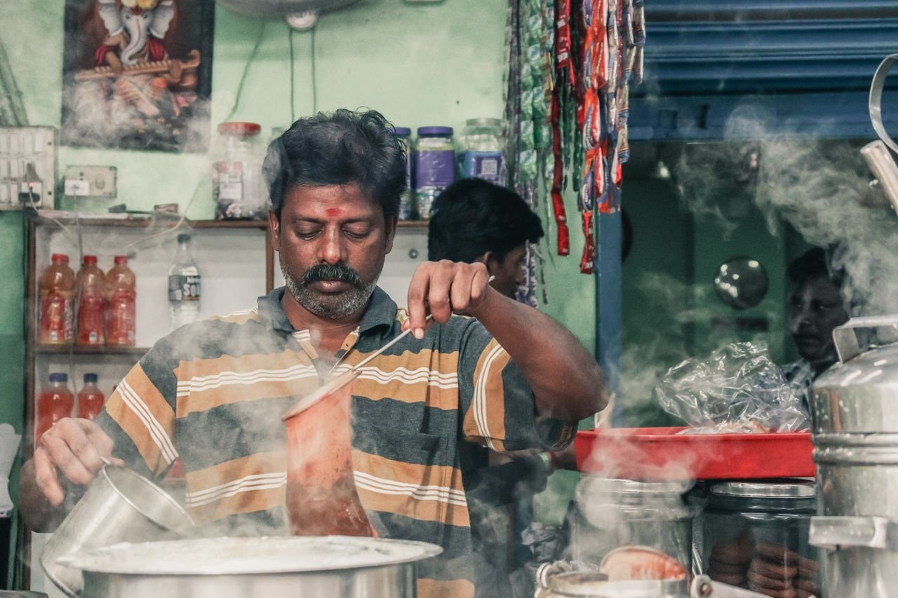 services-03 A man making traditional masala chai in an Indian tea stall, surrounded by steam and kitchen utensils.