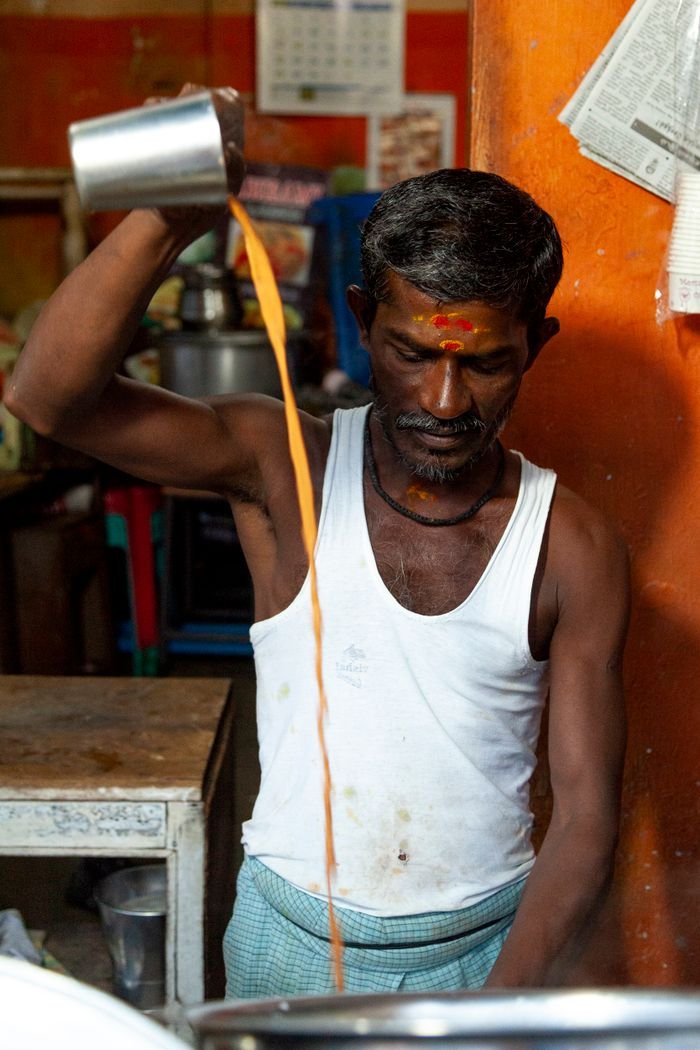 our-story South Asian man in Mahabalipuram pouring chai from a height, displaying traditional method.